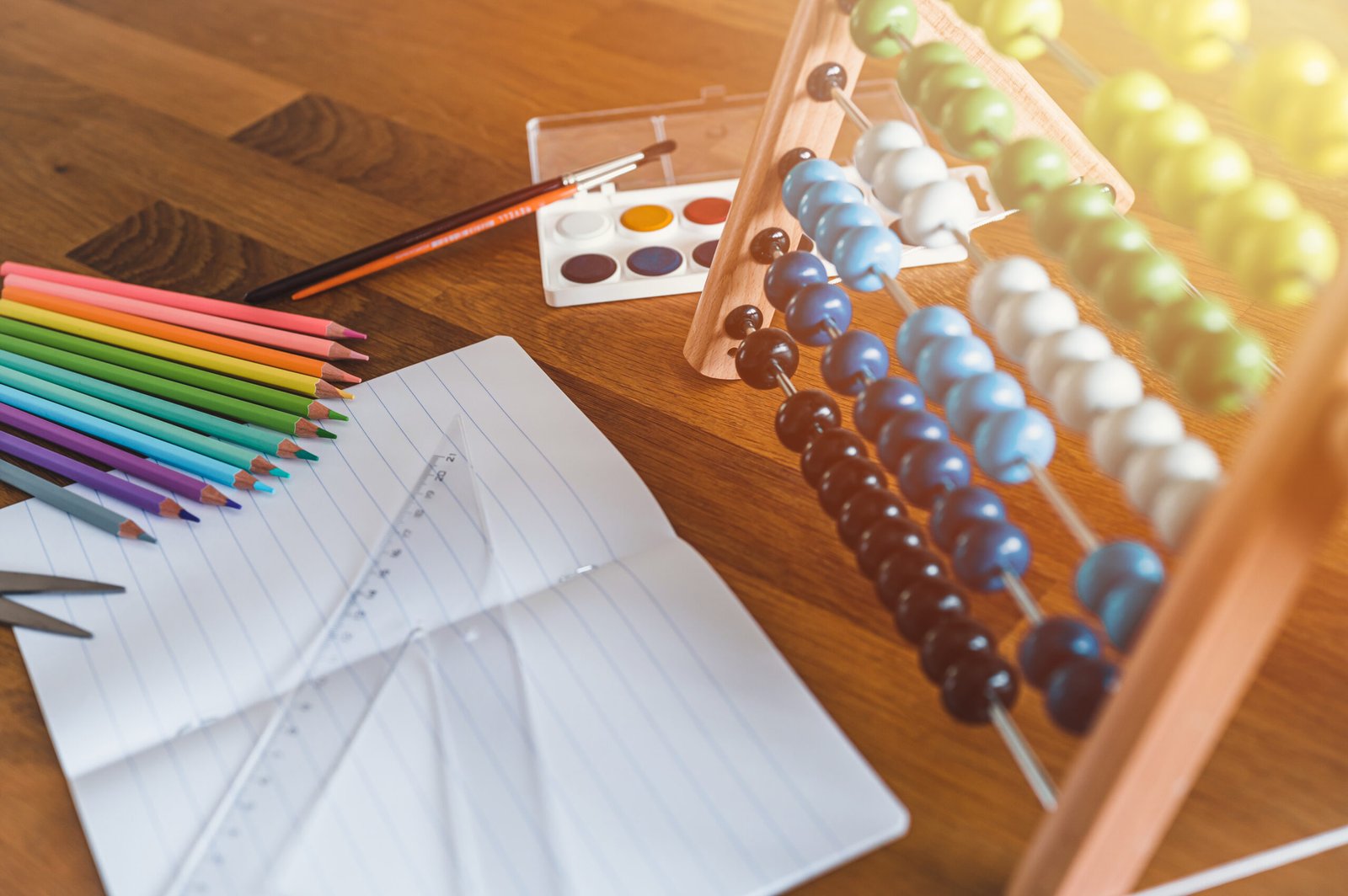 An abacus, colored pencils, paints, and a notebook arranged on a desk